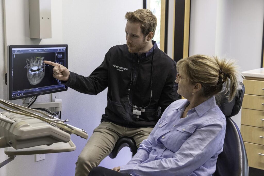 A man stands beside a seated woman, pointing at a computer screen in an office or clinical setting. They appear to be discussing information displayed on the monitor.