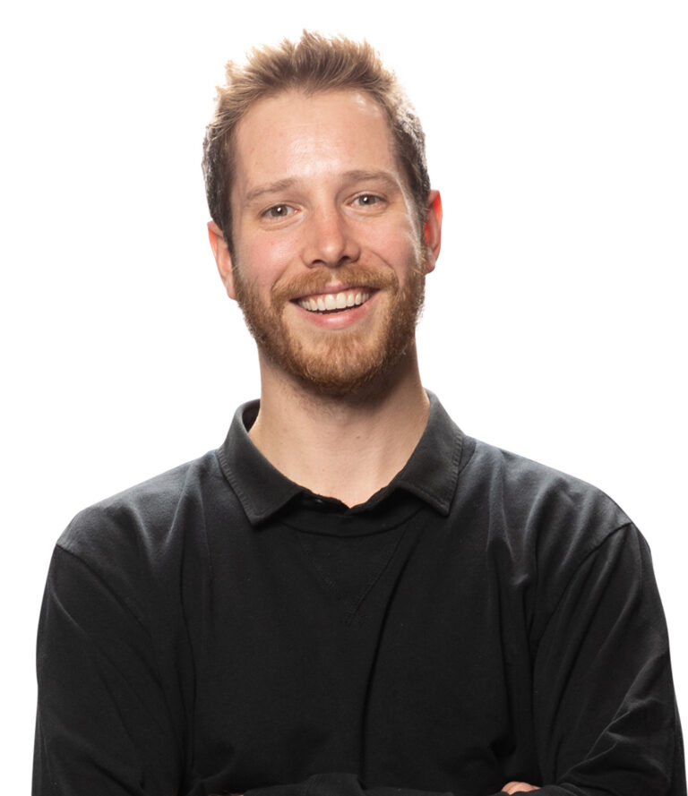 A smiling man wearing a black shirt stands against a plain white background. He faces the camera with a relaxed and confident posture.